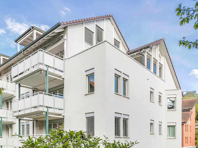 Exterior view of a modern, white apartment building with balconies and a light blue sky.