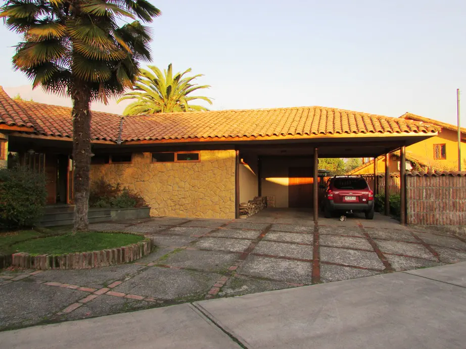 Exterior view of a single-story house with a red tile roof, stone facade, and a carport with a red SUV parked inside. Palm trees are in the front yard.