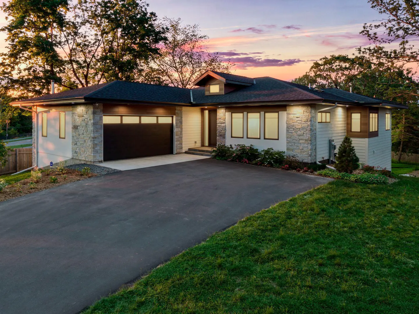 A modern, single-story house with a black garage door, stone accents, and a dark roof at dusk.