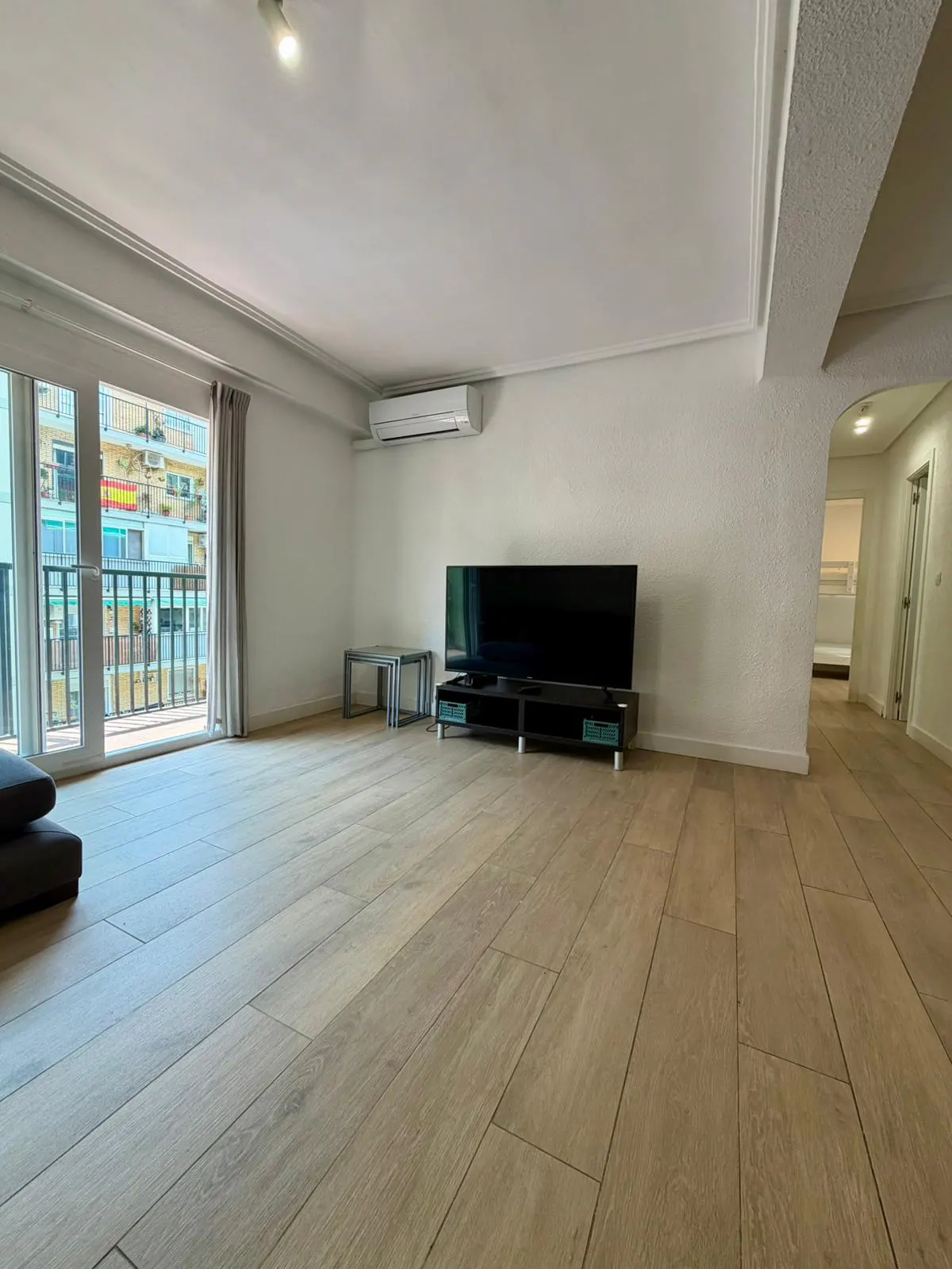Bright living room with wood floors, white walls, and a balcony. A black TV sits on a stand, with a small table nearby.