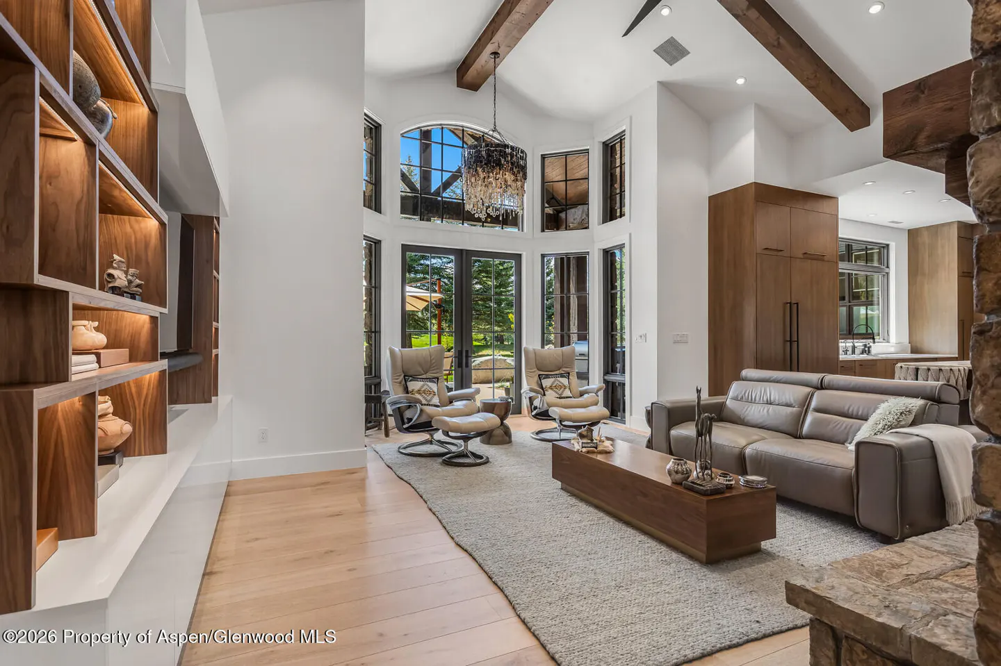 Bright living room with a gray sofa, two chairs, and a wood coffee table on a gray rug. Large windows and a chandelier add elegance.
