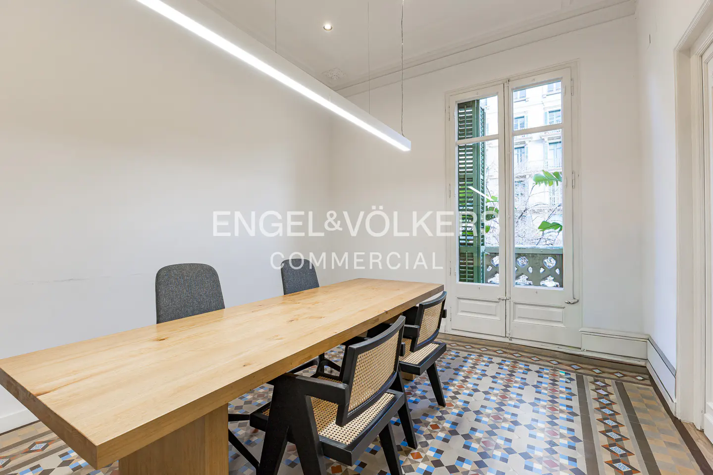 A bright room with a long wooden table and chairs, lit by a linear light fixture. A window with green shutters overlooks a city street.