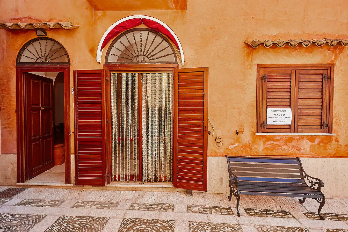 Orange building with open wood doors and shutters. A "Vendesi" sign hangs on the window. A bench sits on the patterned stone sidewalk.