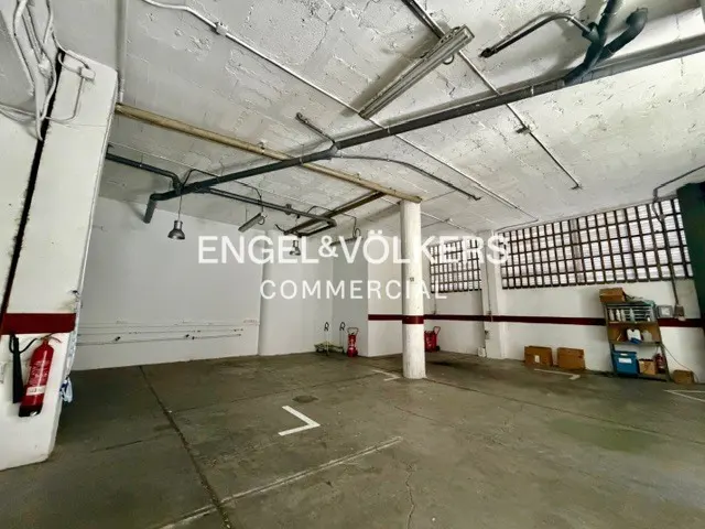A large, empty commercial space with concrete floors, white walls, and exposed pipes and lighting on the ceiling. Fire extinguishers are visible.