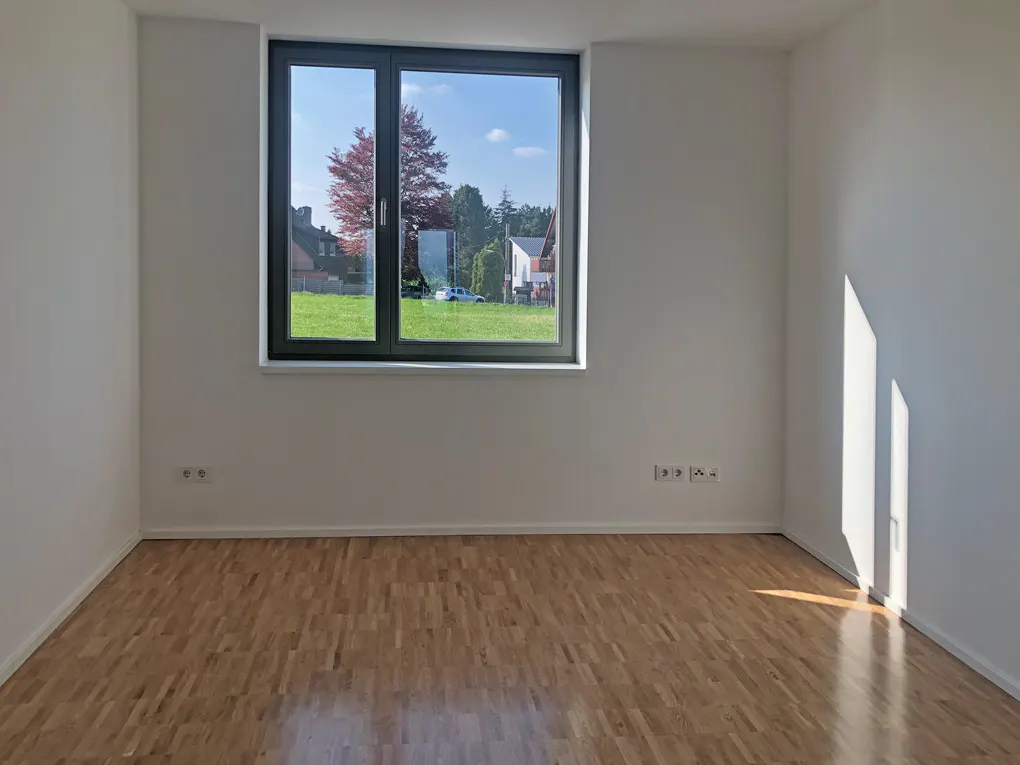 Empty room with light wood floor, white walls, and a large window showing a green lawn and trees.