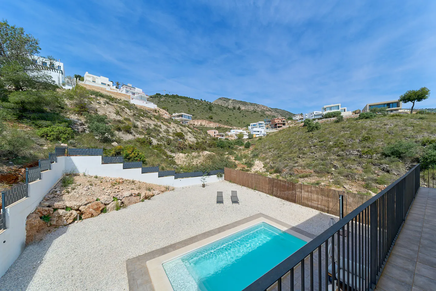 A rectangular pool with turquoise water, two lounge chairs, and hillside homes under a blue sky.