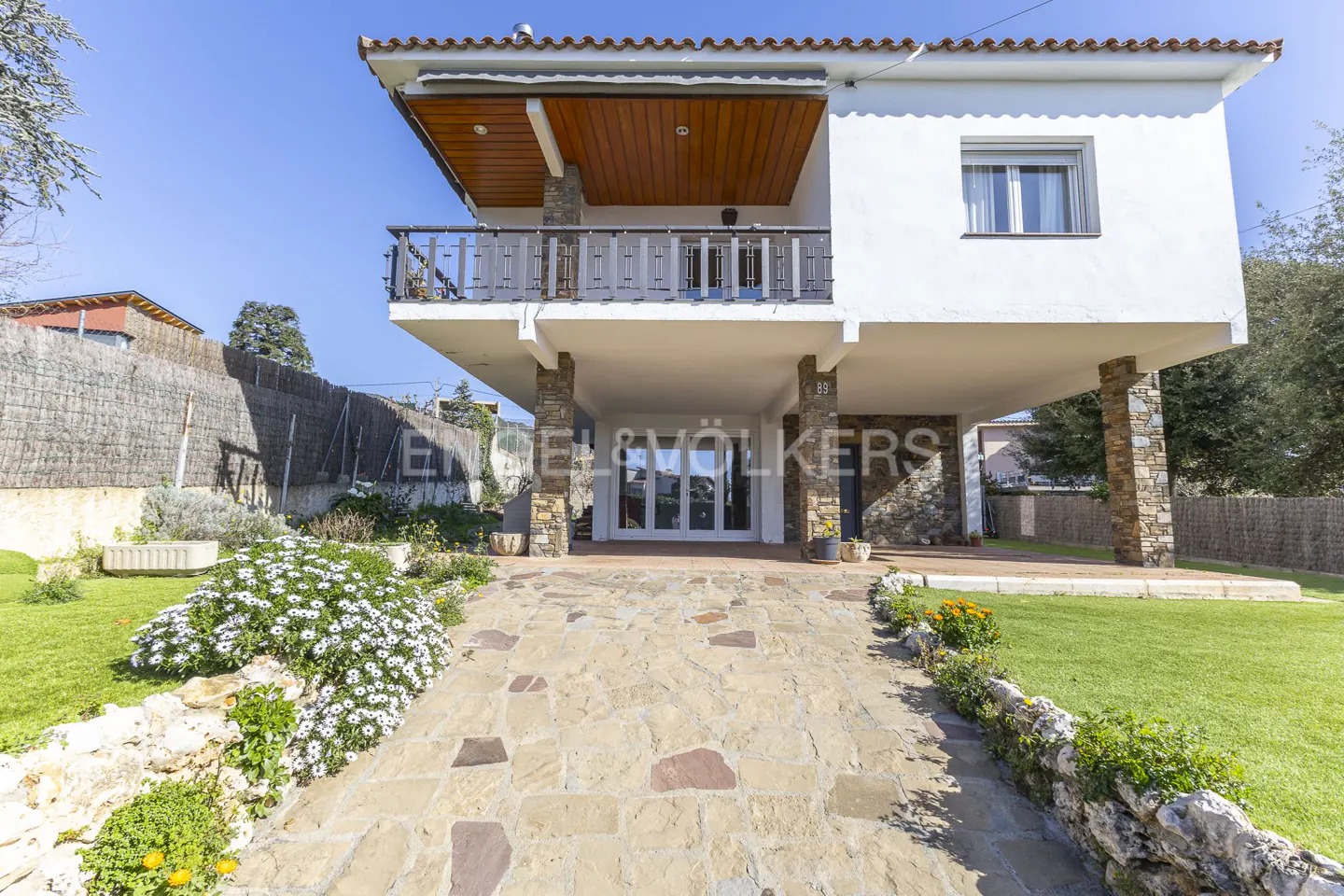 Two-story white house with stone pillars and a stone walkway leading to the entrance. Green lawn and flowers surround the house.