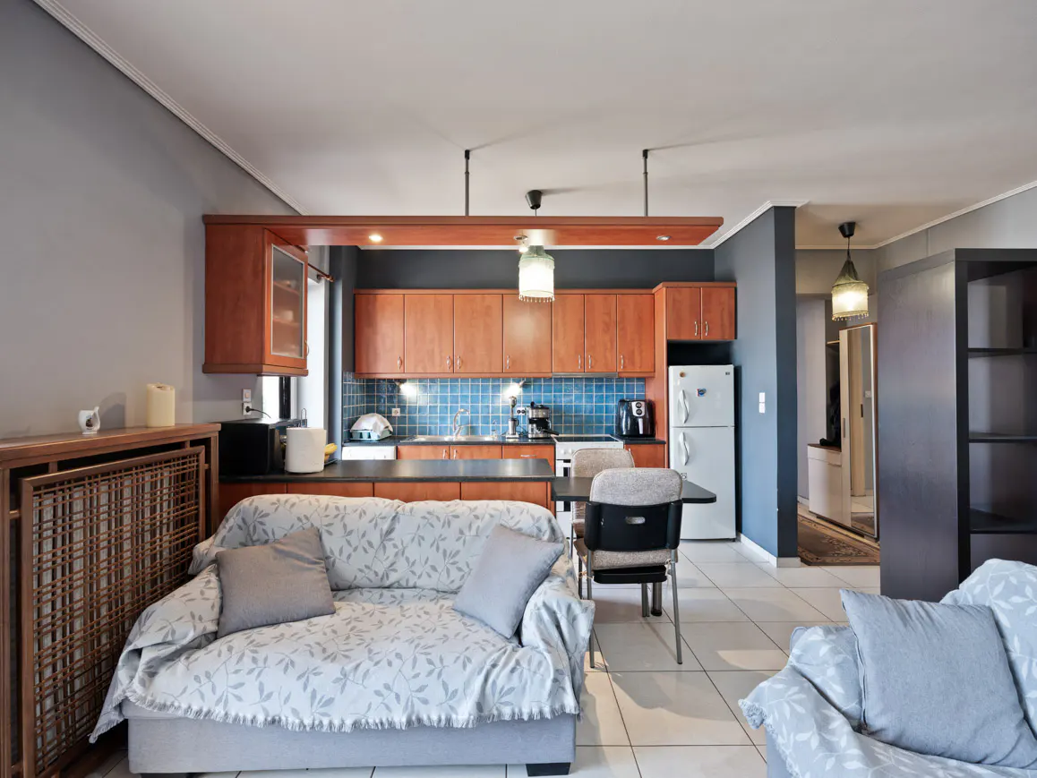 A bright apartment interior shows a living room with a kitchen. The kitchen has wood cabinets and blue tile. A gray sofa is in the foreground.