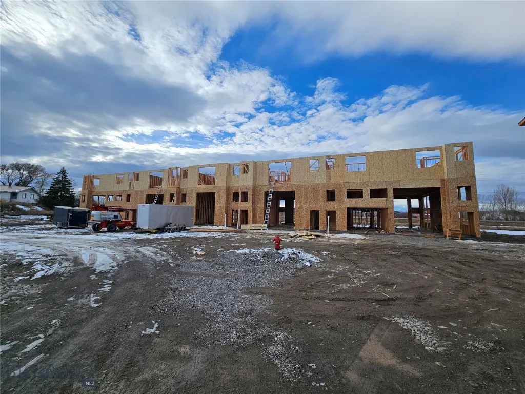 A partially constructed wood-framed building stands on a dirt lot with snow patches under a cloudy sky.