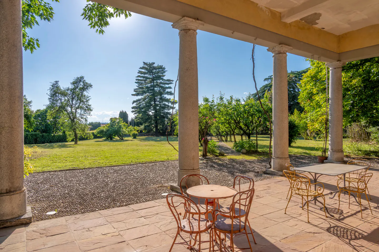 Covered patio with stone columns, featuring outdoor seating and a view of a green lawn and trees on a sunny day.