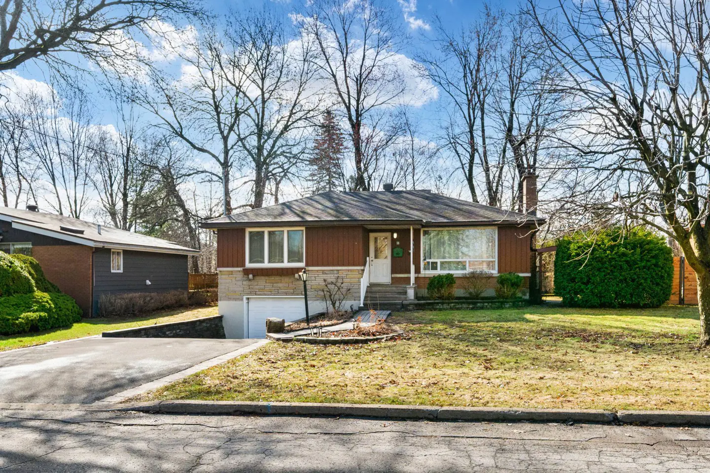 A one-story house with brown siding and a stone foundation, a white door, and a driveway leading to a garage.