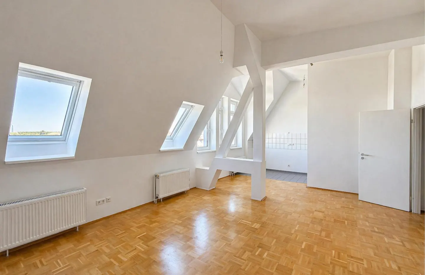 Bright, empty attic apartment with white walls, wood floors, skylights, and white support beams.