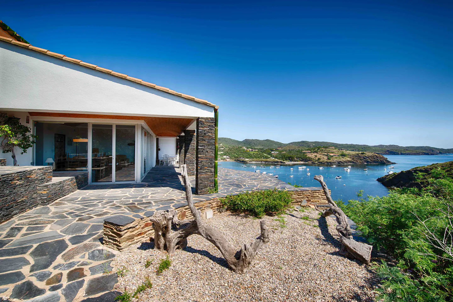 Modern villa with stone patio and driftwood overlooking a blue bay with boats under a clear sky.