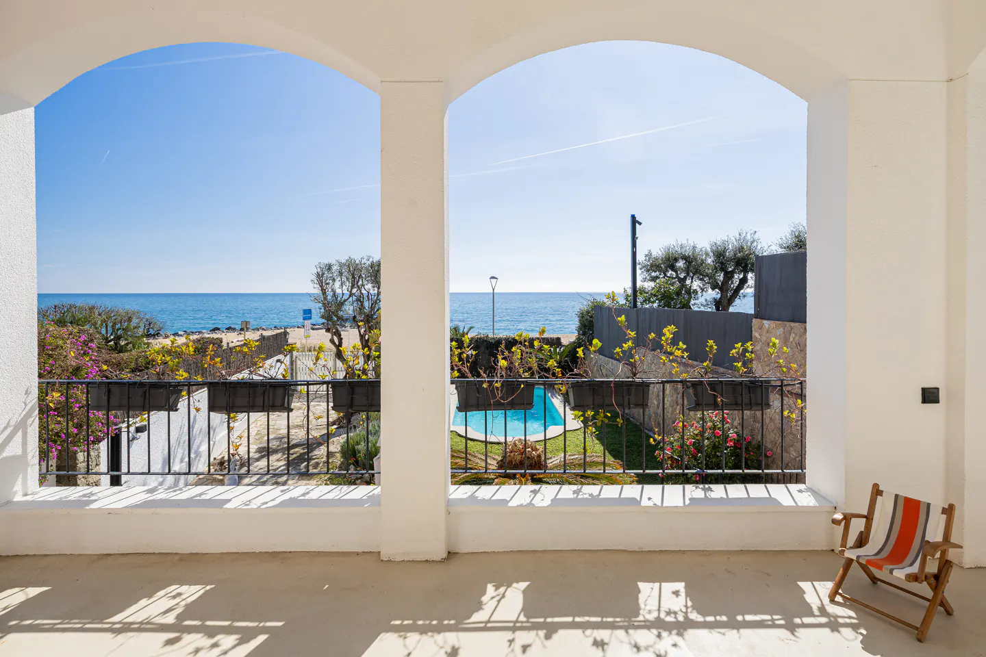View from a white arched terrace overlooking a swimming pool, garden, and the blue sea under a clear sky.