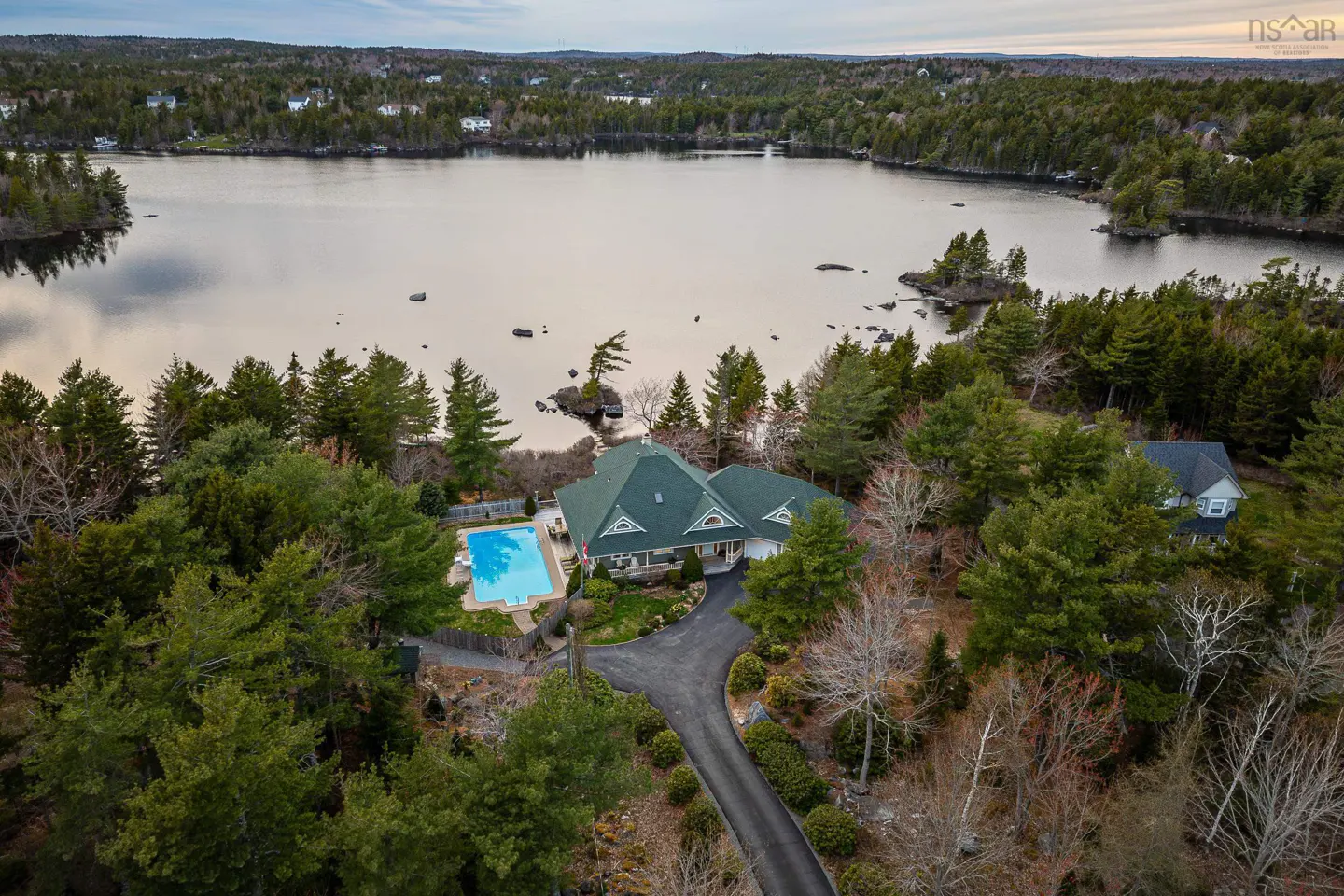 Aerial view of a waterfront home with a green roof, a pool, and a long driveway surrounded by trees. A lake is visible in the background.