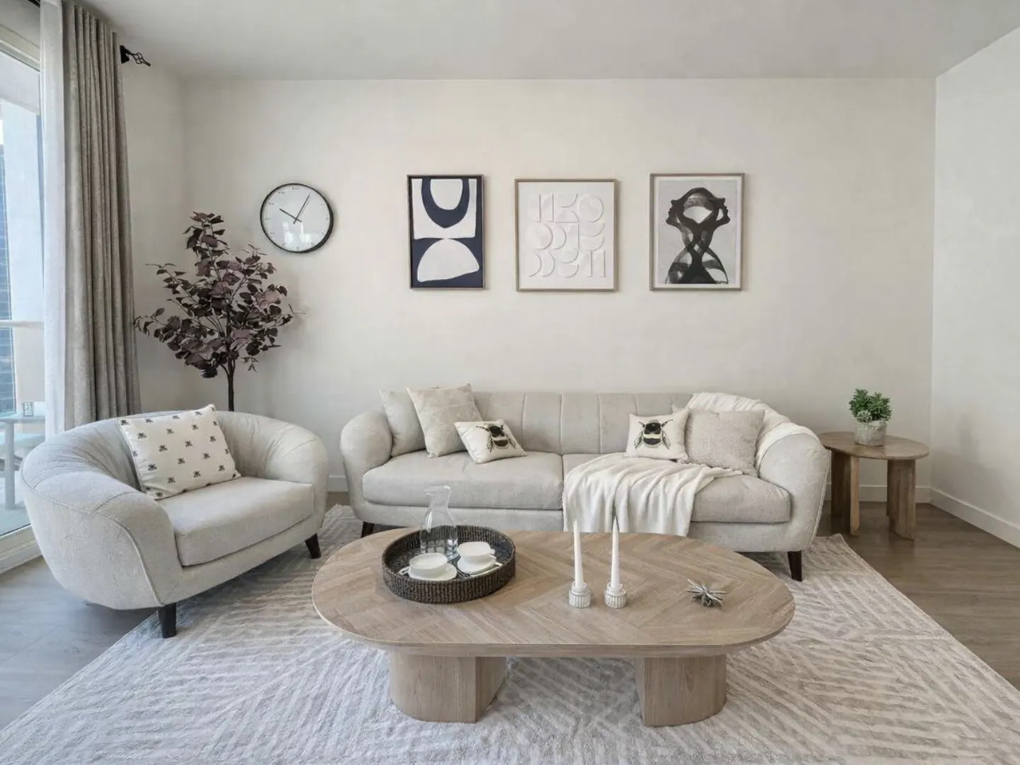 Modern living room with a beige sofa and armchair, oval wooden coffee table, patterned rug, and three abstract wall prints.