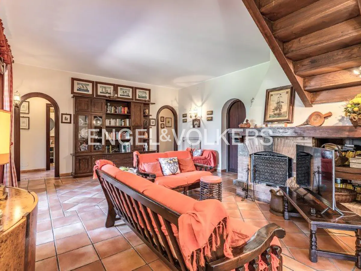 Living room with terracotta tile floor, orange sofas, stone fireplace, and wooden staircase. Arched doorways and built-in bookcase add character.