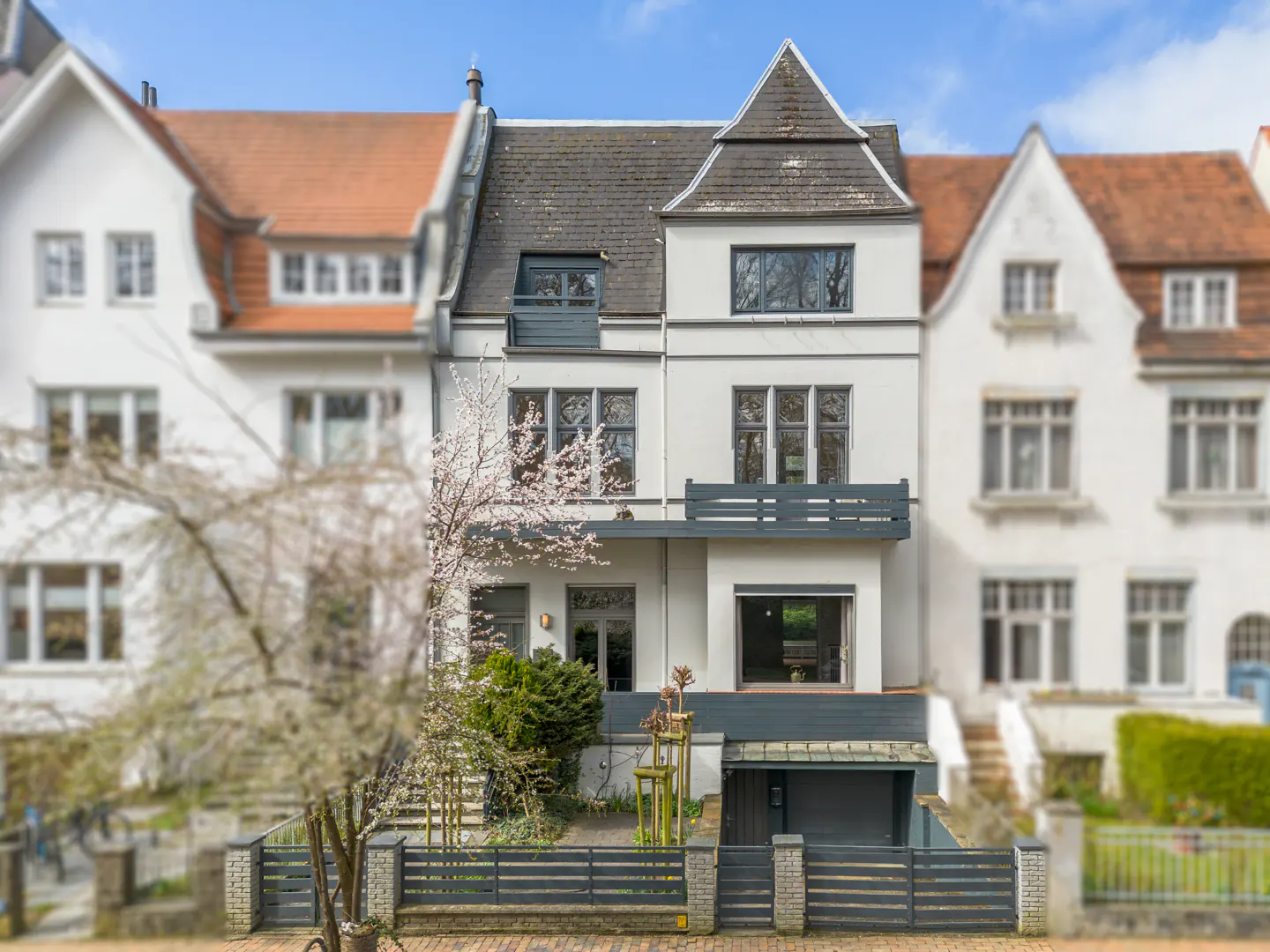 A three-story white house with a gray roof and a small balcony is centered in the frame. Other houses are blurred in the background.