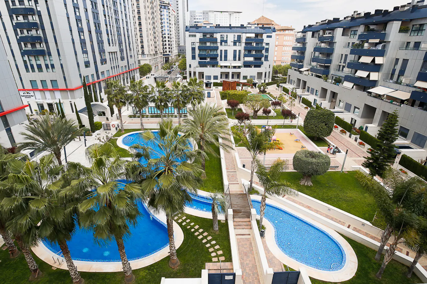Aerial view of a residential complex with pools, palm trees, a playground, and modern apartment buildings.
