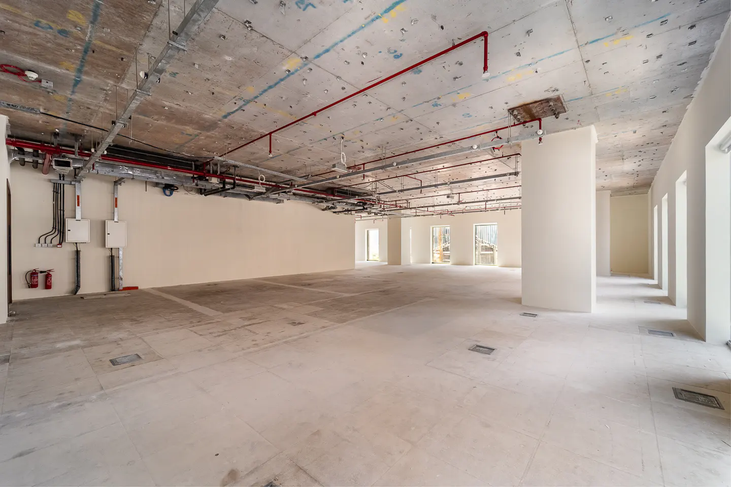 Unfinished office space with concrete floors and ceiling. Red pipes and metal conduits run along the ceiling. White walls and window frames.