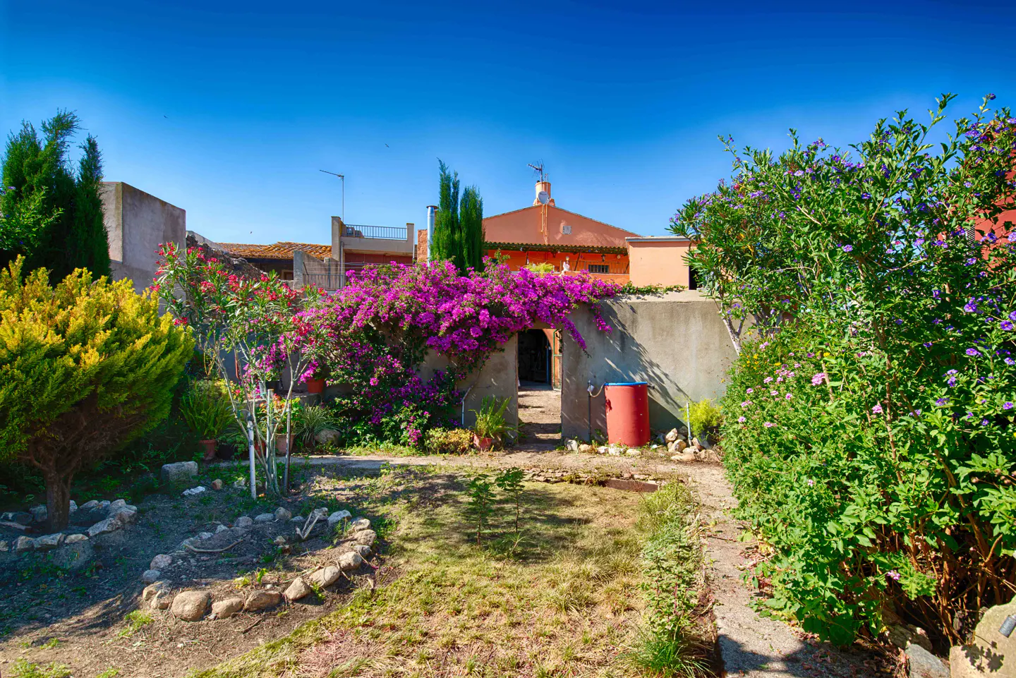 A garden with a stone archway covered in purple bougainvillea leads to a house under a clear blue sky.