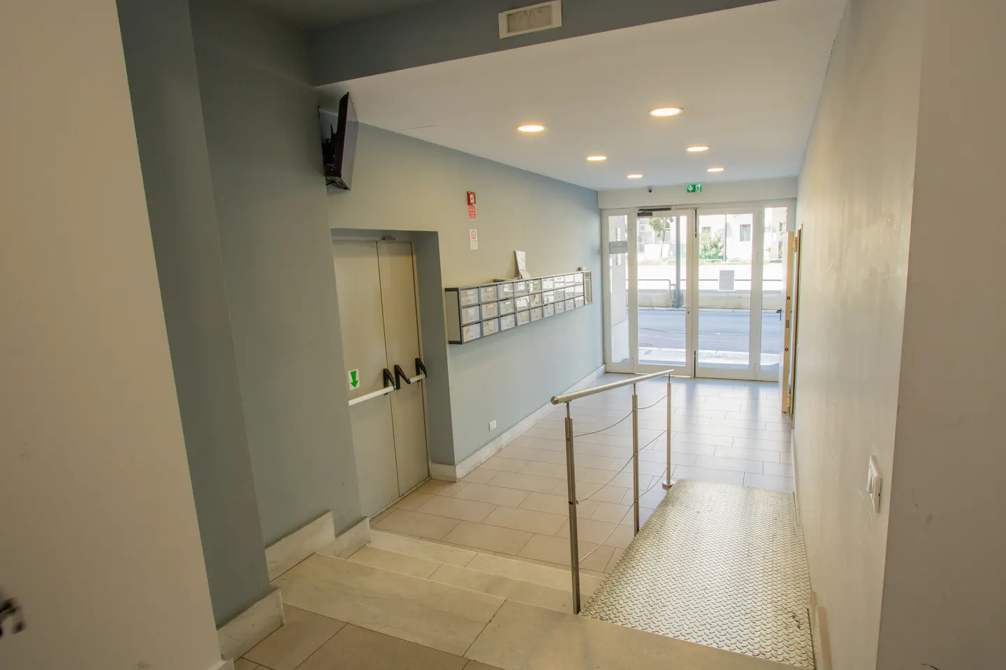 Indoor view of a building entrance with an elevator, mailboxes, and glass doors leading outside. The walls are painted in light gray.