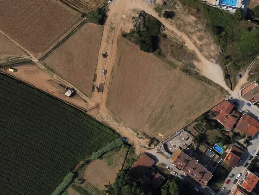 Aerial view of brown fields, green crops, and red-roofed houses. A dirt road runs through the fields.