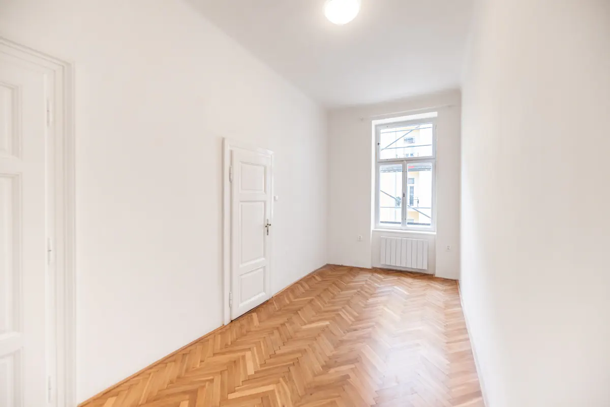 Bright, empty room with white walls, herringbone wood floor, and a window with a radiator below. Two white doors are visible.