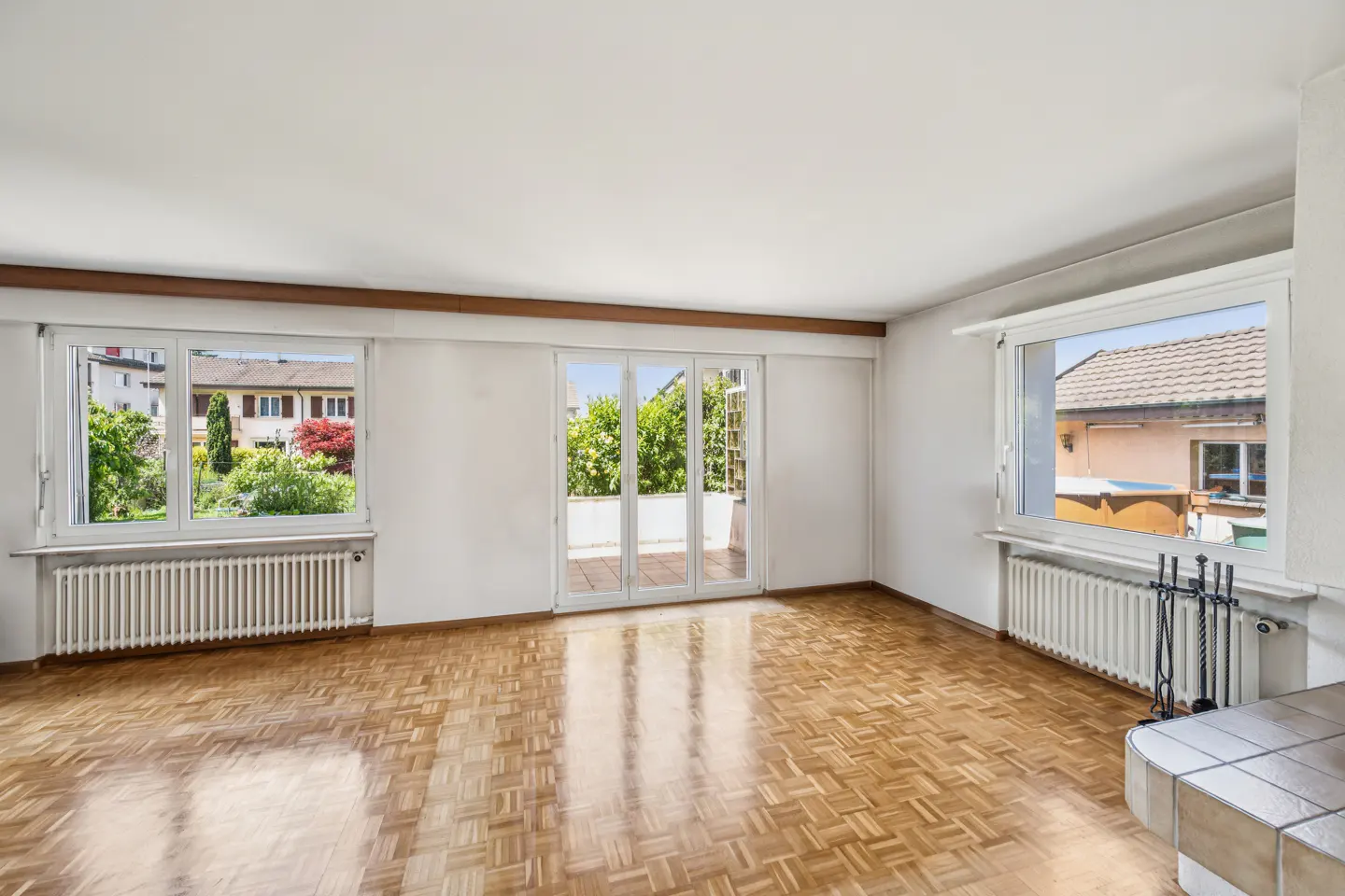 Bright, empty room with parquet floors, white walls, and three windows. The center window is a door to a balcony. Radiators are under the windows.