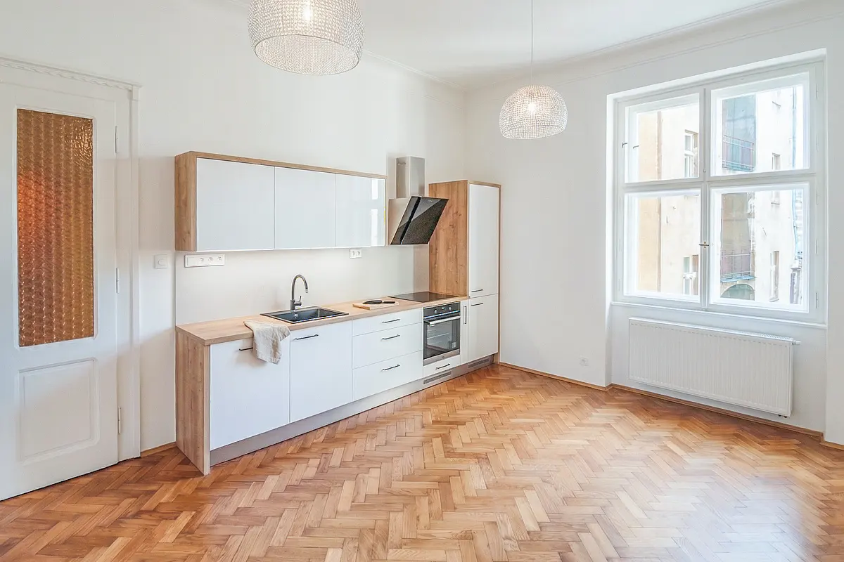 Bright, modern kitchen with white cabinets, wood countertops, and herringbone wood floors. Large window and decorative pendant lights.