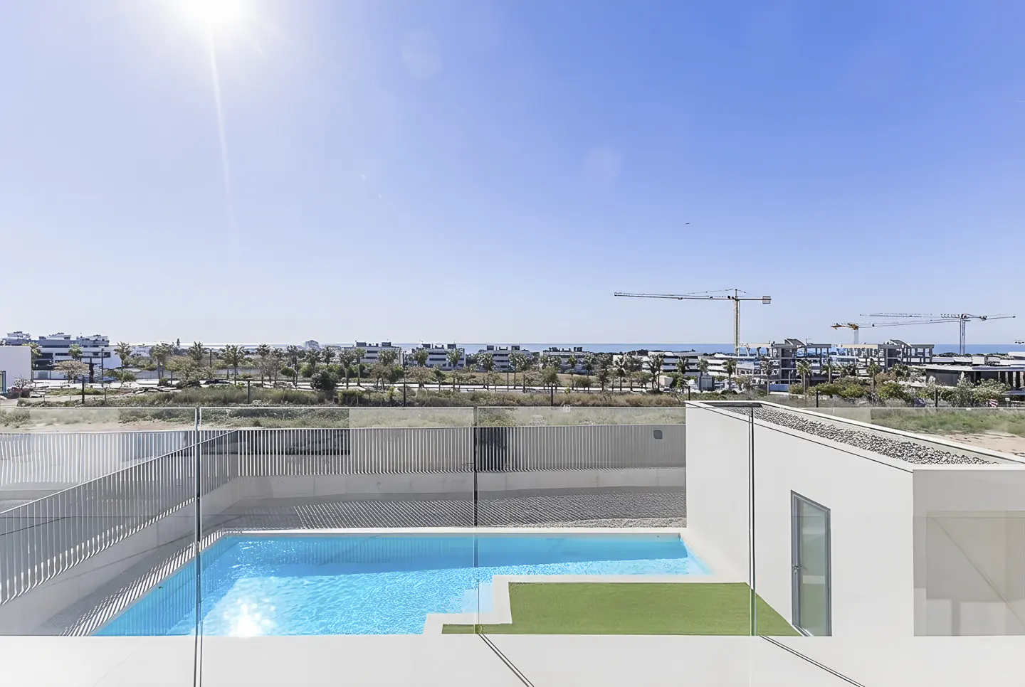View of a blue pool with a green patch, a white building, and a city skyline under a clear blue sky.