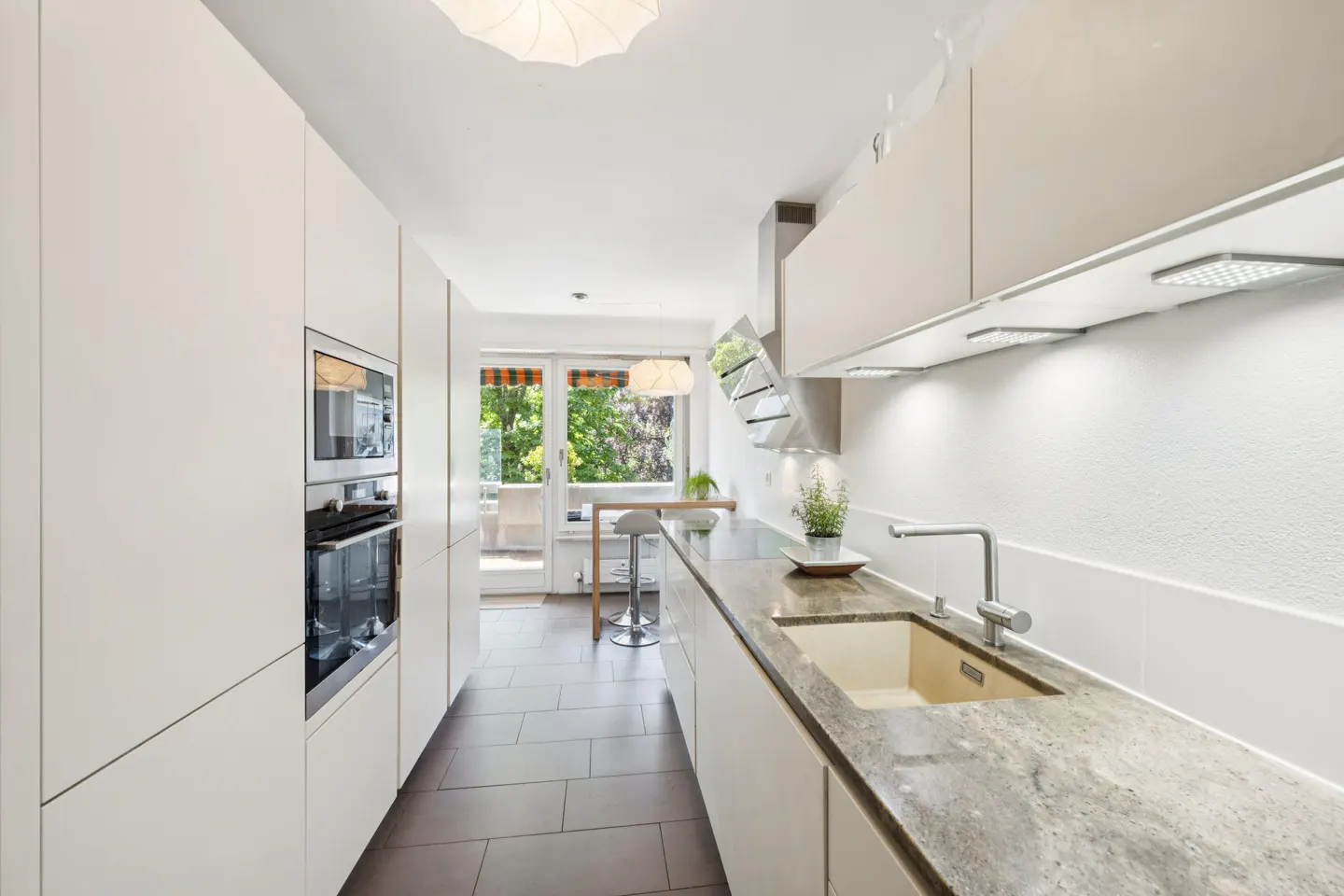 Bright, modern kitchen with white cabinets, granite countertops, and stainless steel appliances. A window overlooks a balcony with green trees.