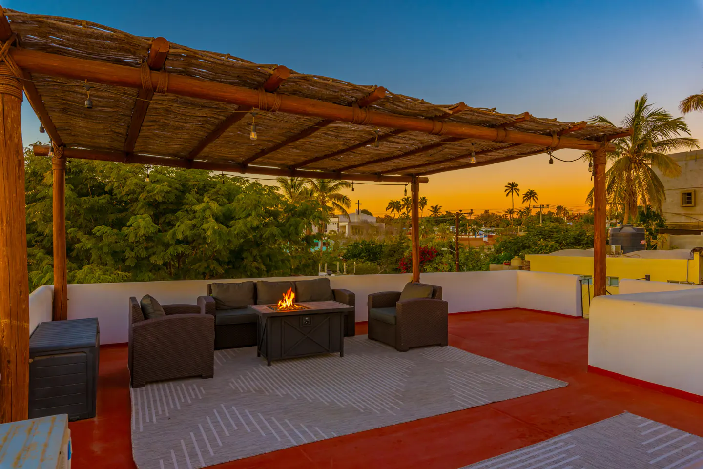 Rooftop patio with a thatched pergola, outdoor furniture, and a fire pit. Sunset sky and palm trees in the background.