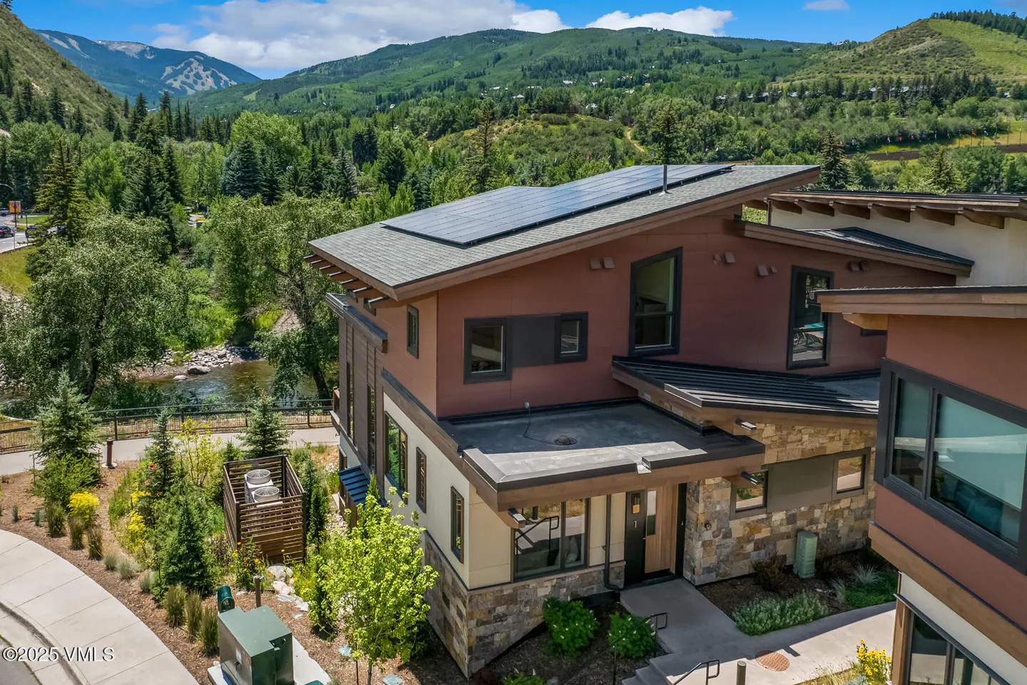 Exterior view of a modern brown and tan home with solar panels, set against a backdrop of green mountains and trees.