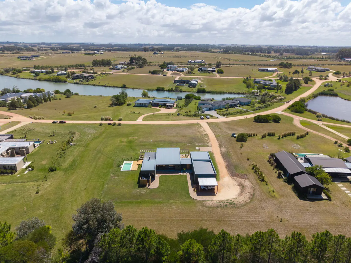 Aerial view of a modern, gray-roofed house with a pool, set in a green, rural landscape with a river and scattered homes.
