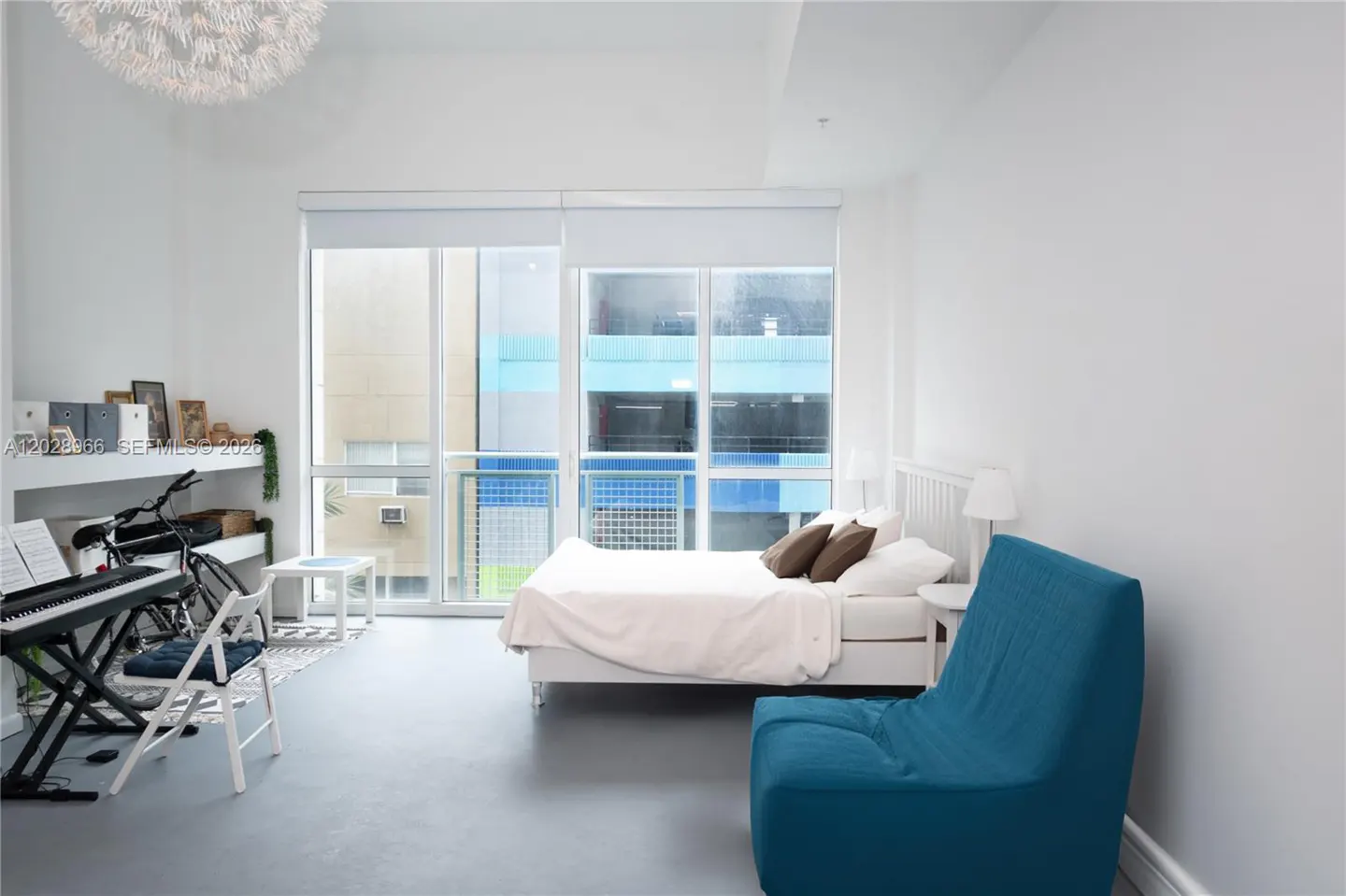 Modern studio apartment with white walls, a bed, a blue armchair, a keyboard on a stand, and large windows with city views.