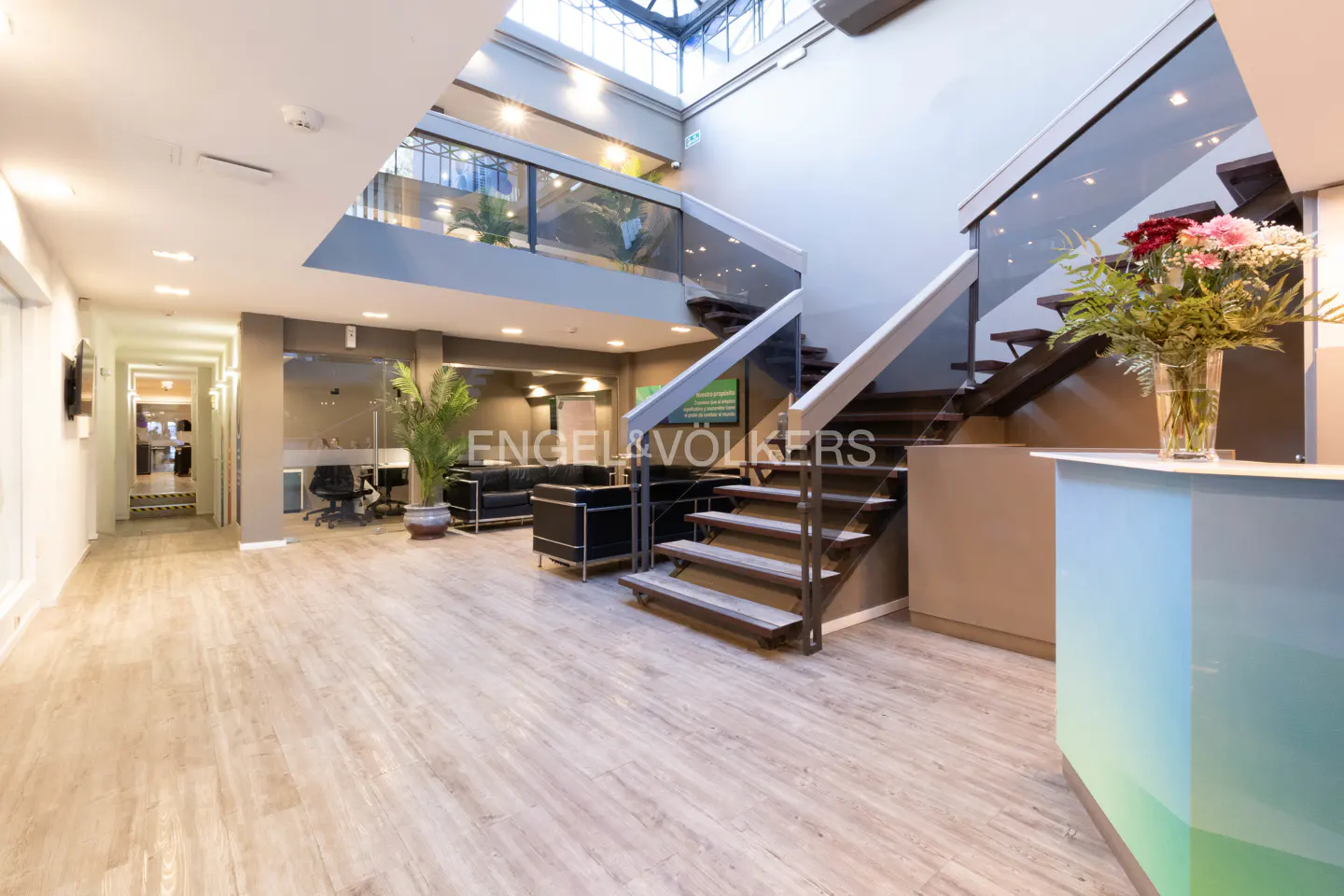 A real estate office interior with wood floors, black sofas, and a staircase. A glass vase with flowers sits on the reception desk.