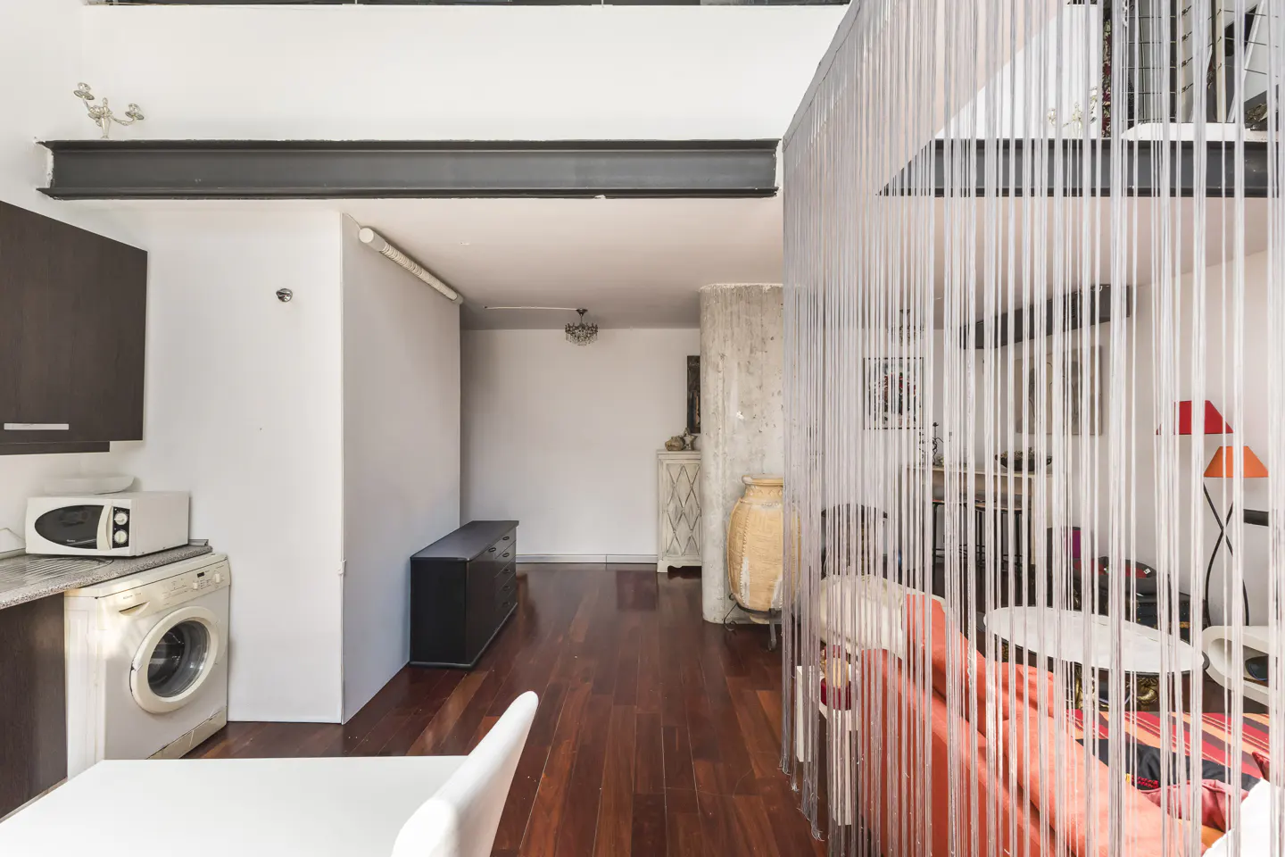 Interior view of a modern apartment with a kitchen area, living room, and loft space, featuring hardwood floors and white walls.