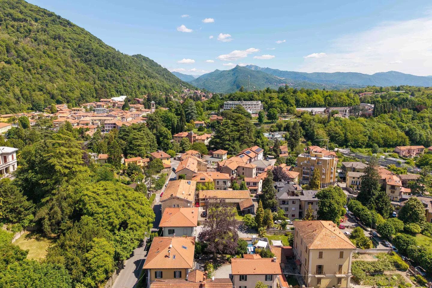 Aerial view of a European town with red-tiled roofs, surrounded by green trees and mountains under a blue sky.