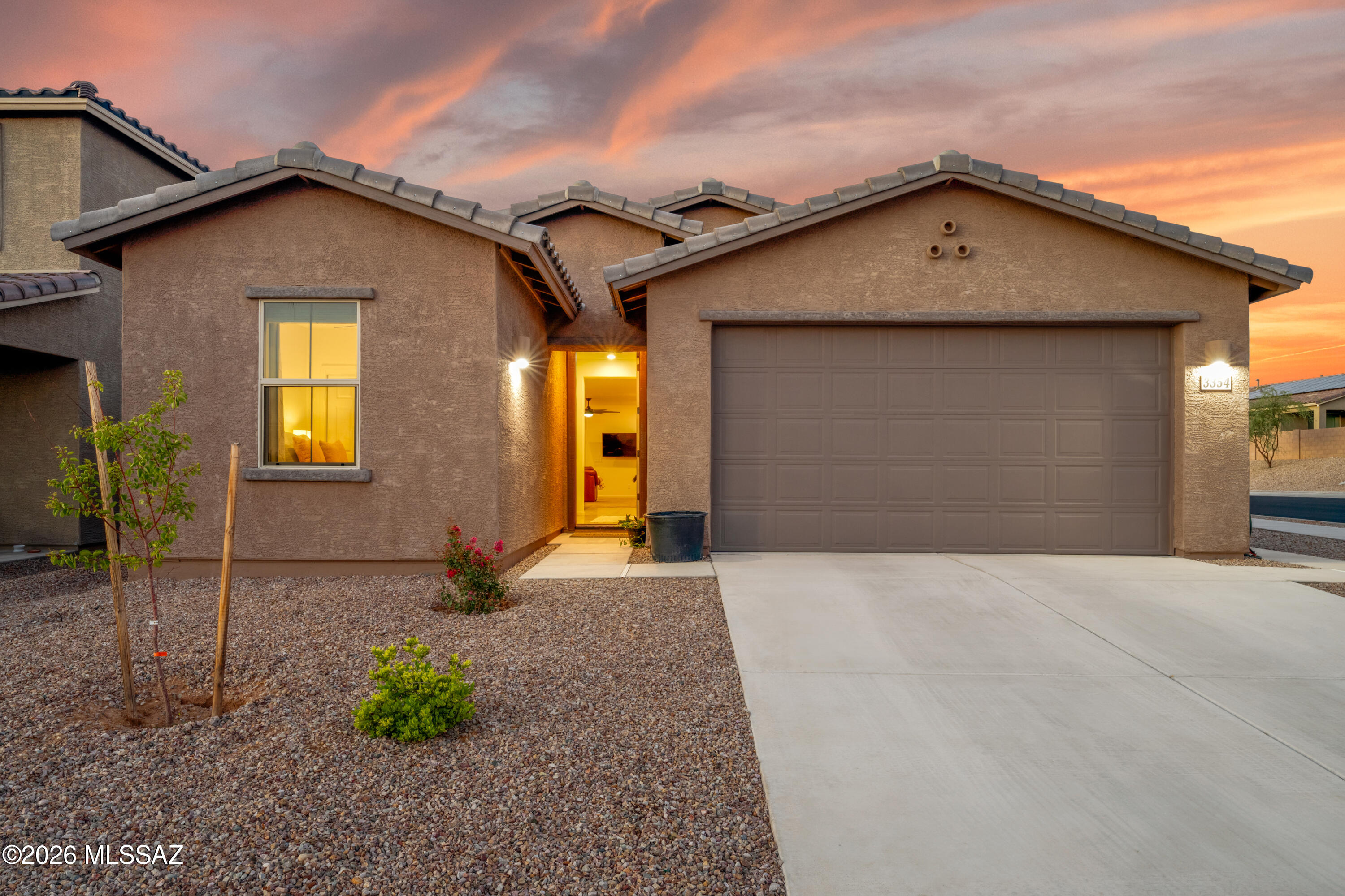 Single-story tan stucco home with a two-car garage and desert landscaping under a vibrant orange sunset sky.