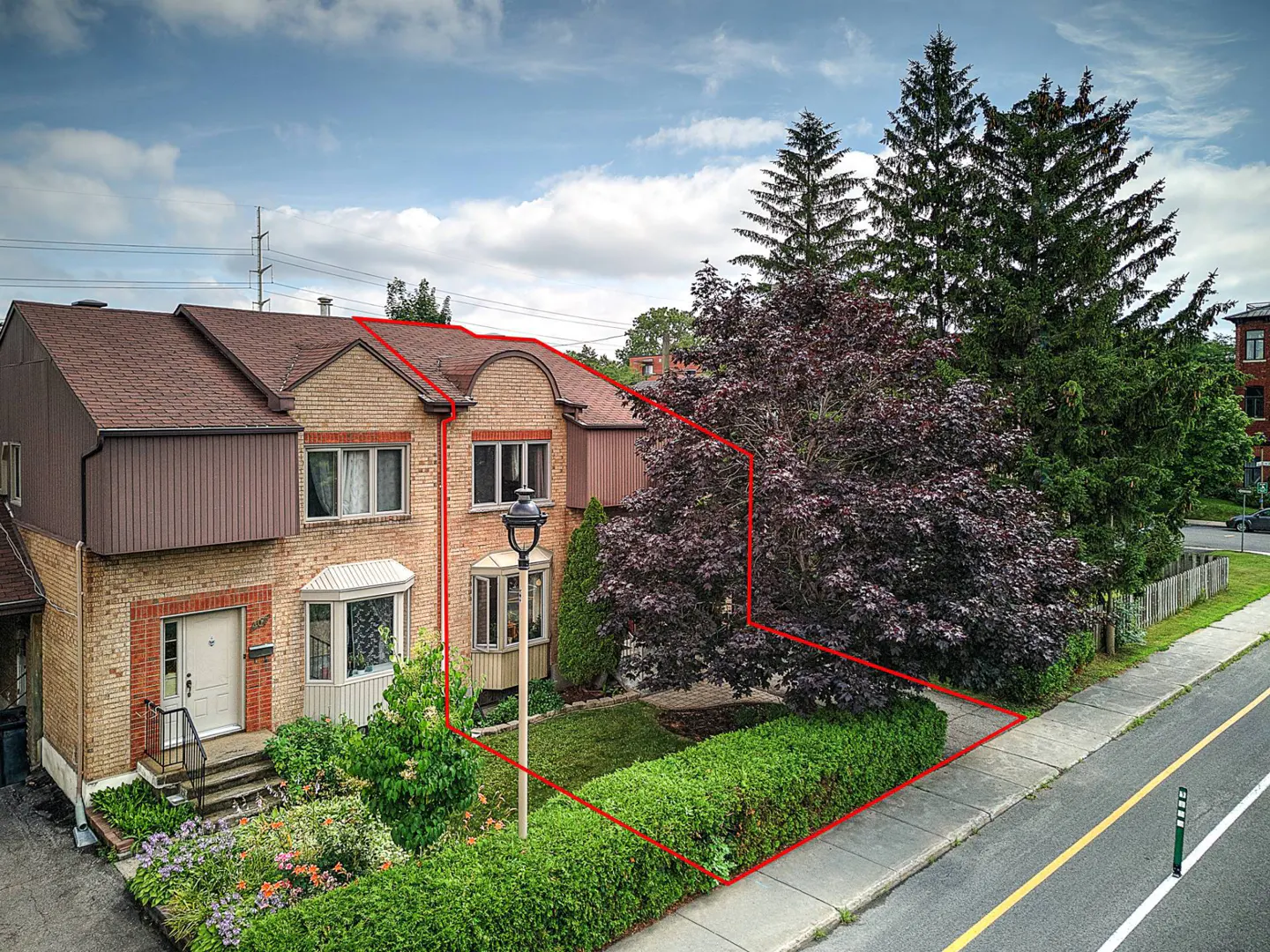 Brick townhouse with a brown roof, outlined in red, has a landscaped front yard with flowers and a large tree.
