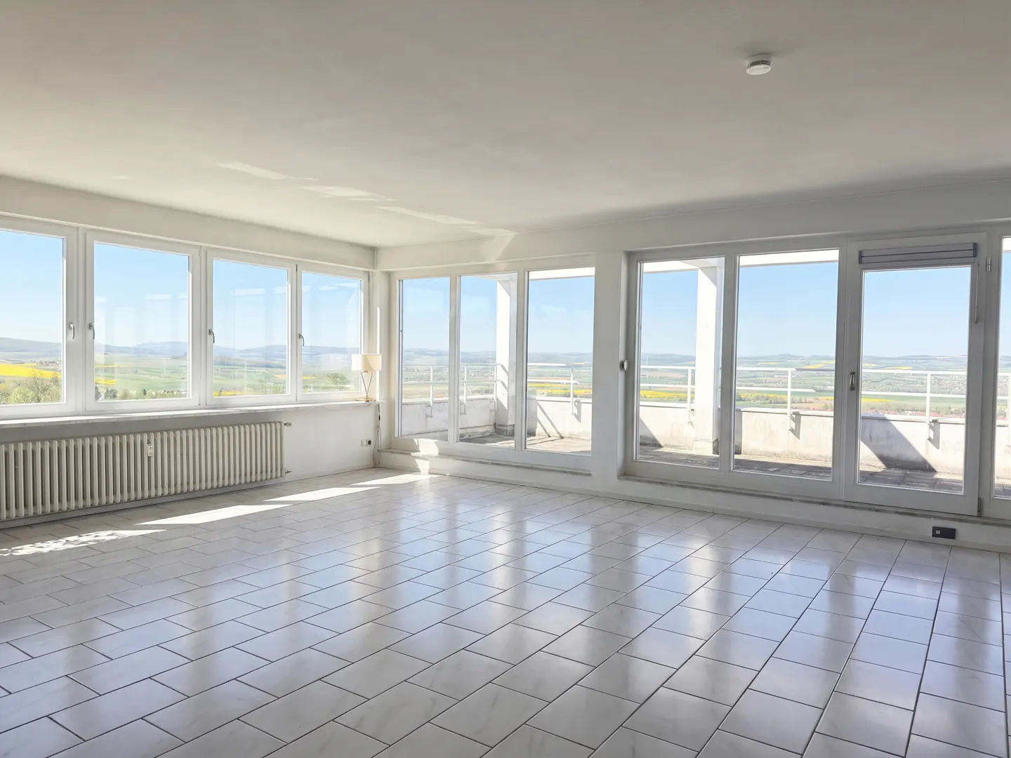 Bright, empty room with white tile floor and large windows showing a landscape view. A radiator sits under the windows.