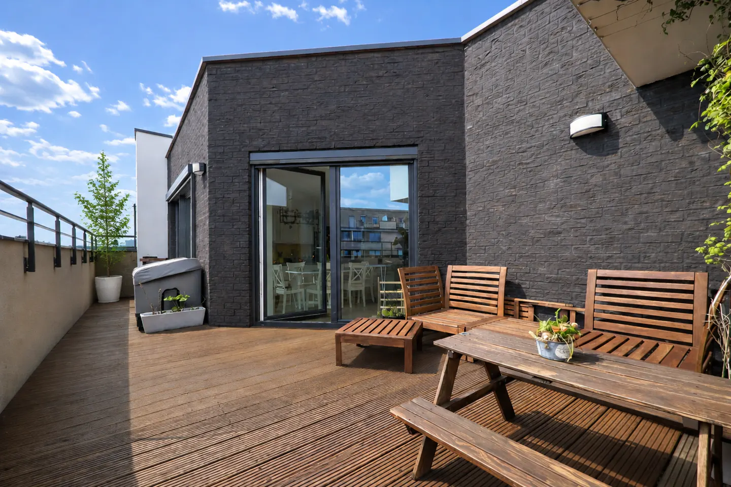 A modern rooftop patio with a wooden deck, picnic table, chairs, and a black brick building. Sliding glass doors lead inside.