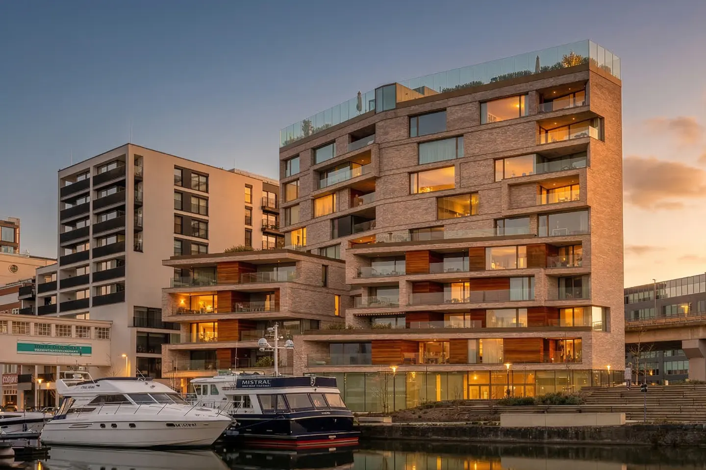 Modern brick apartment building with balconies and large windows overlooking a marina with docked yachts at sunset.