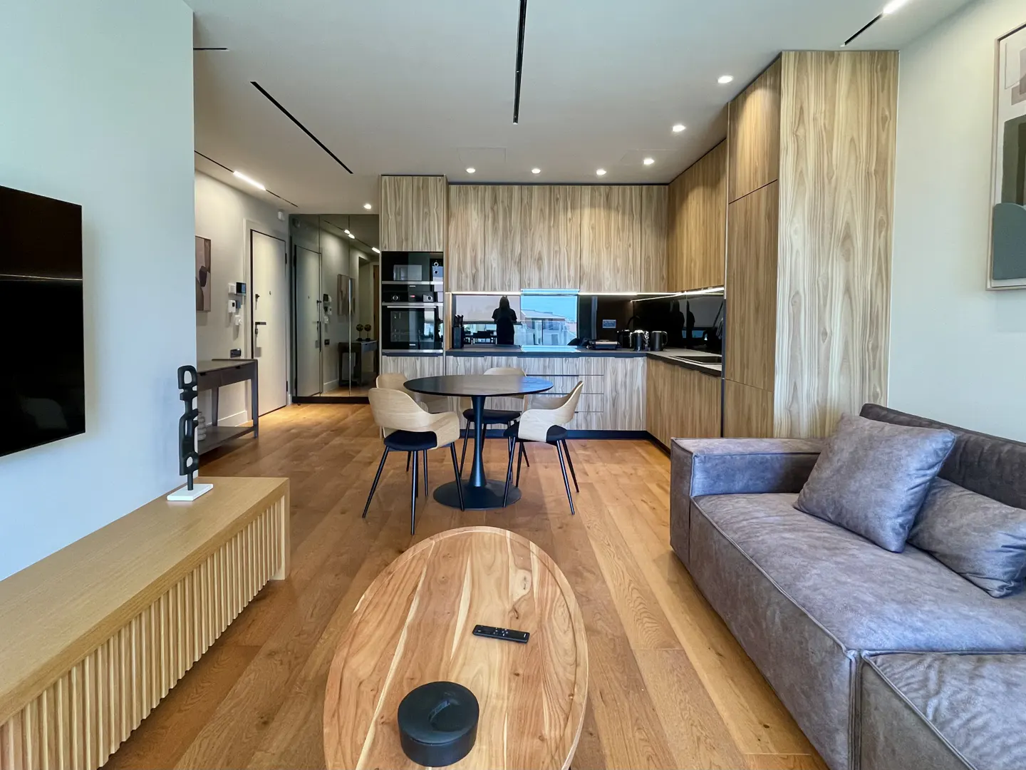 A modern apartment interior with wood floors, a gray sofa, a black round table with four chairs, and a wood-grain kitchen.