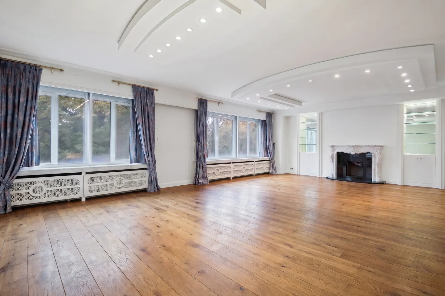 Bright, empty living room with hardwood floors, white walls, and blue patterned curtains framing large windows. A marble fireplace and built-in shelves add architectural detail.