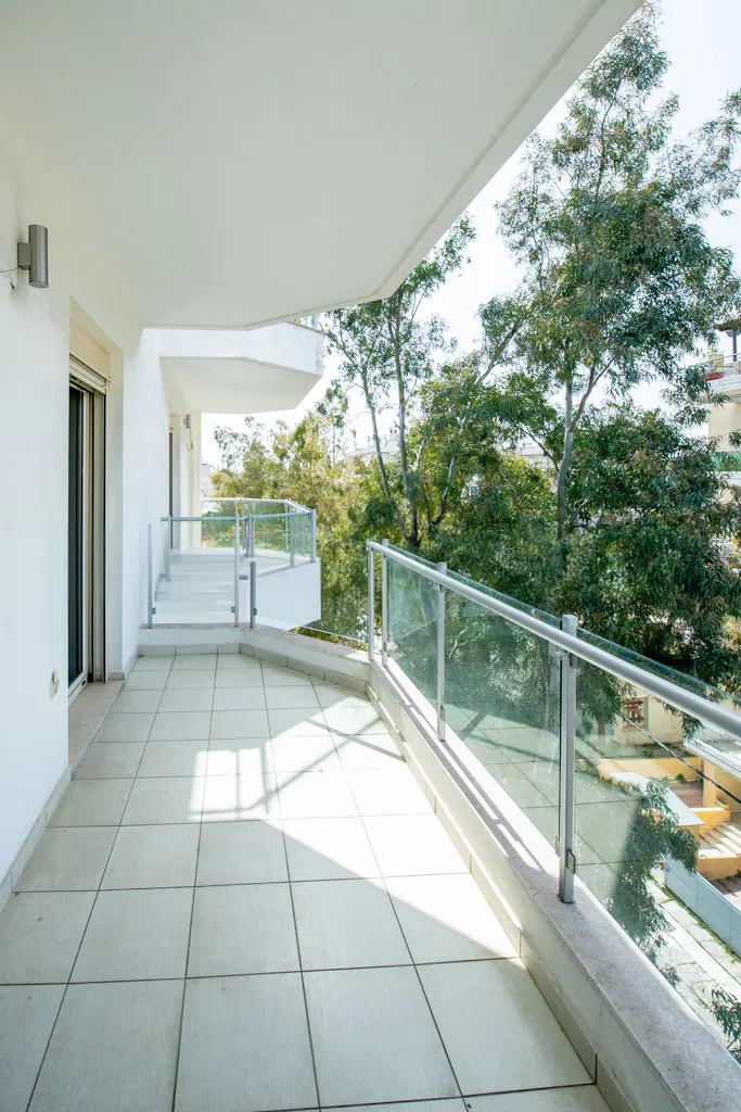 A modern white balcony with glass railings and tiled floor overlooks lush green trees.