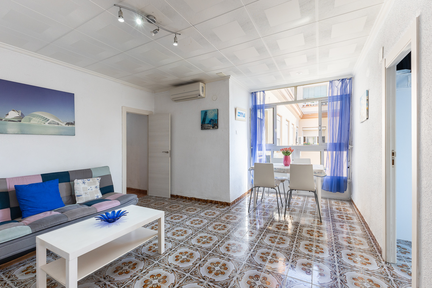 Living room with a patchwork sofa, white coffee table, and dining area with blue curtains and patterned tile floors.