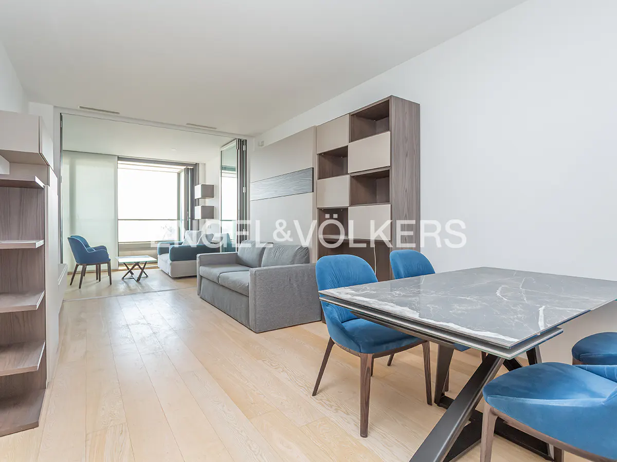 Bright living room with light wood floors, gray sofa, blue chairs, and a marble-top dining table. Balcony visible through sliding glass doors.