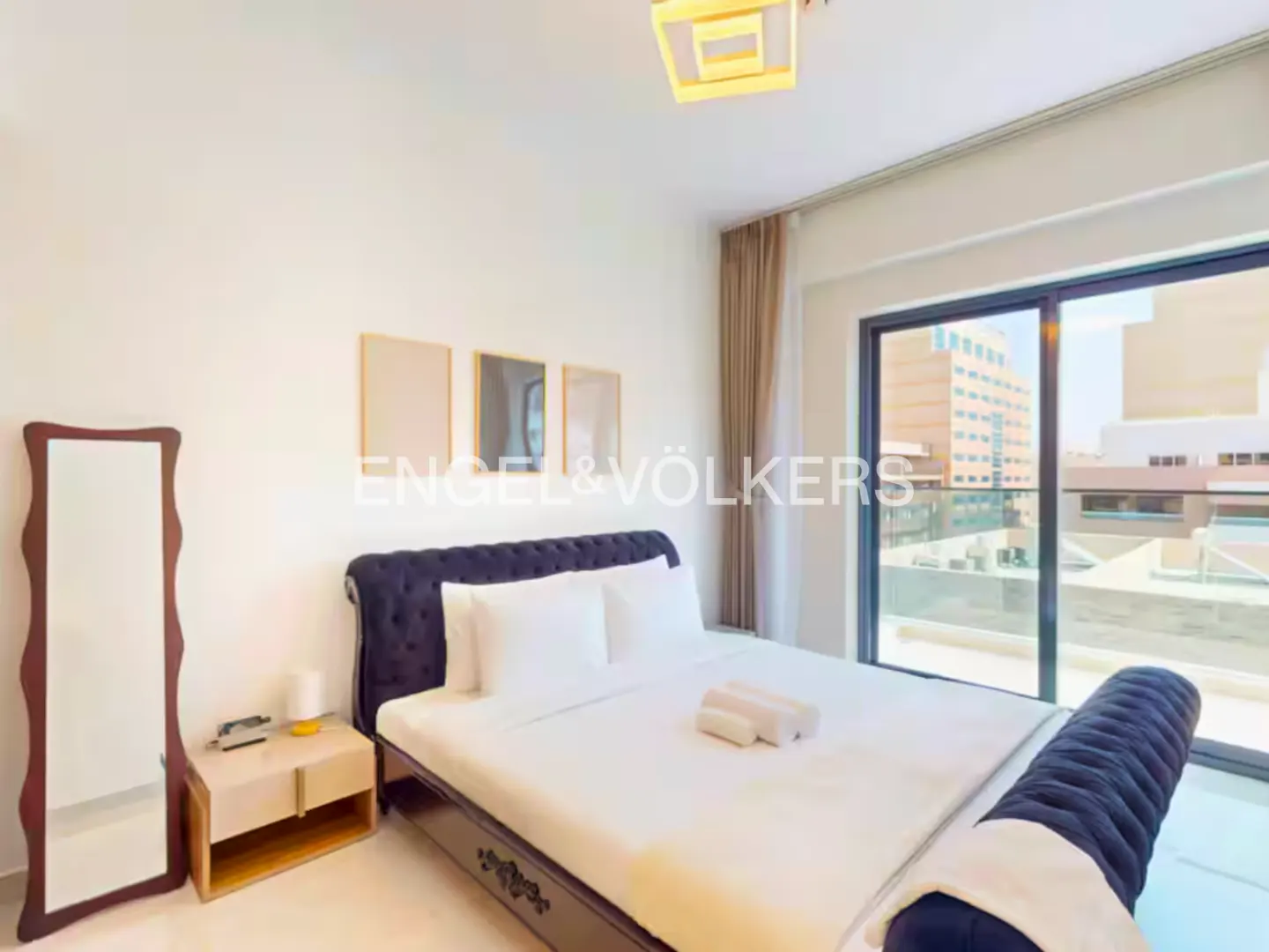 Bedroom with a navy tufted bed, white linens, and a city view from a large window. A full-length mirror and nightstand are visible.